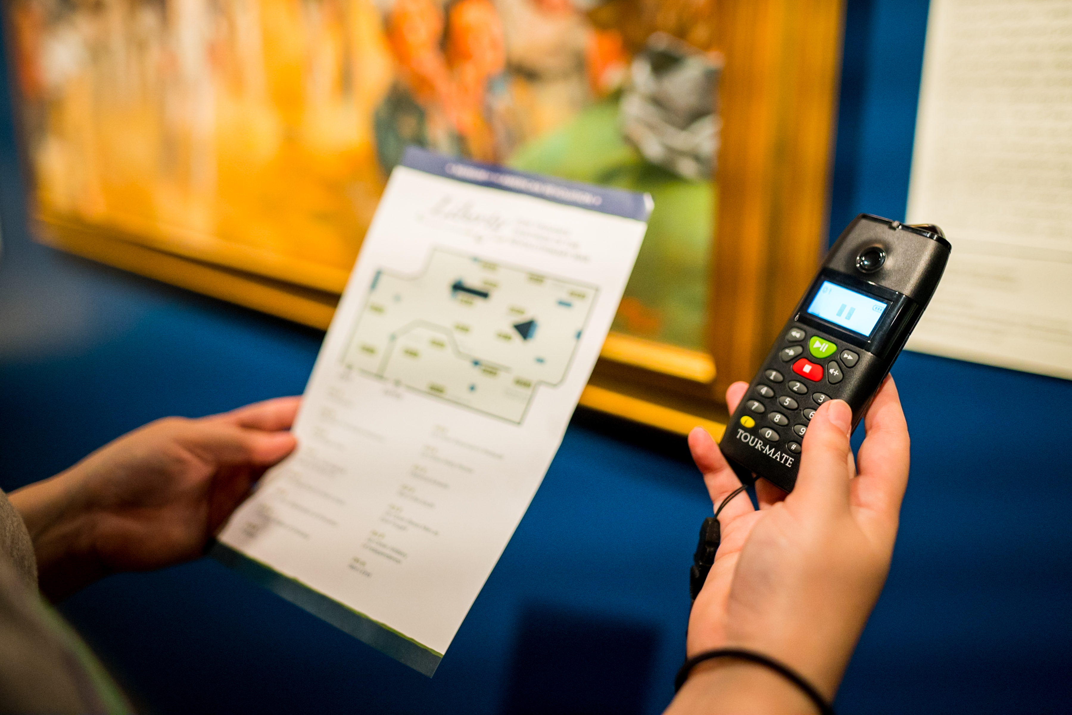 A guest holds a Museum-issued audio tour device in their right hand and the printed audio tour guide in their left.