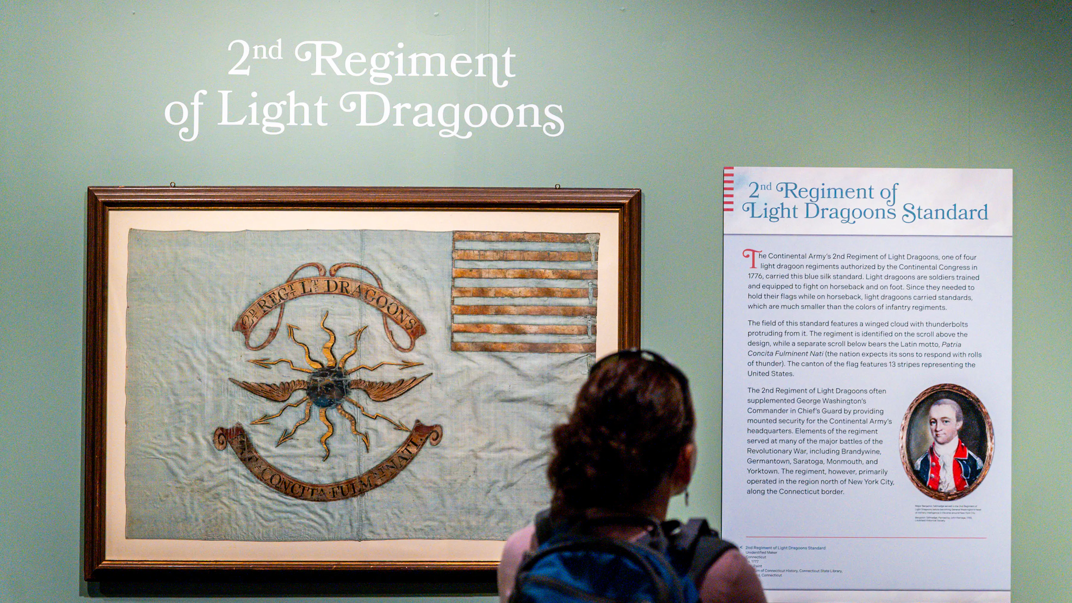 This image shows a woman looking at a light blue Revolutionary War flag and a Museum panel hung over a green wall