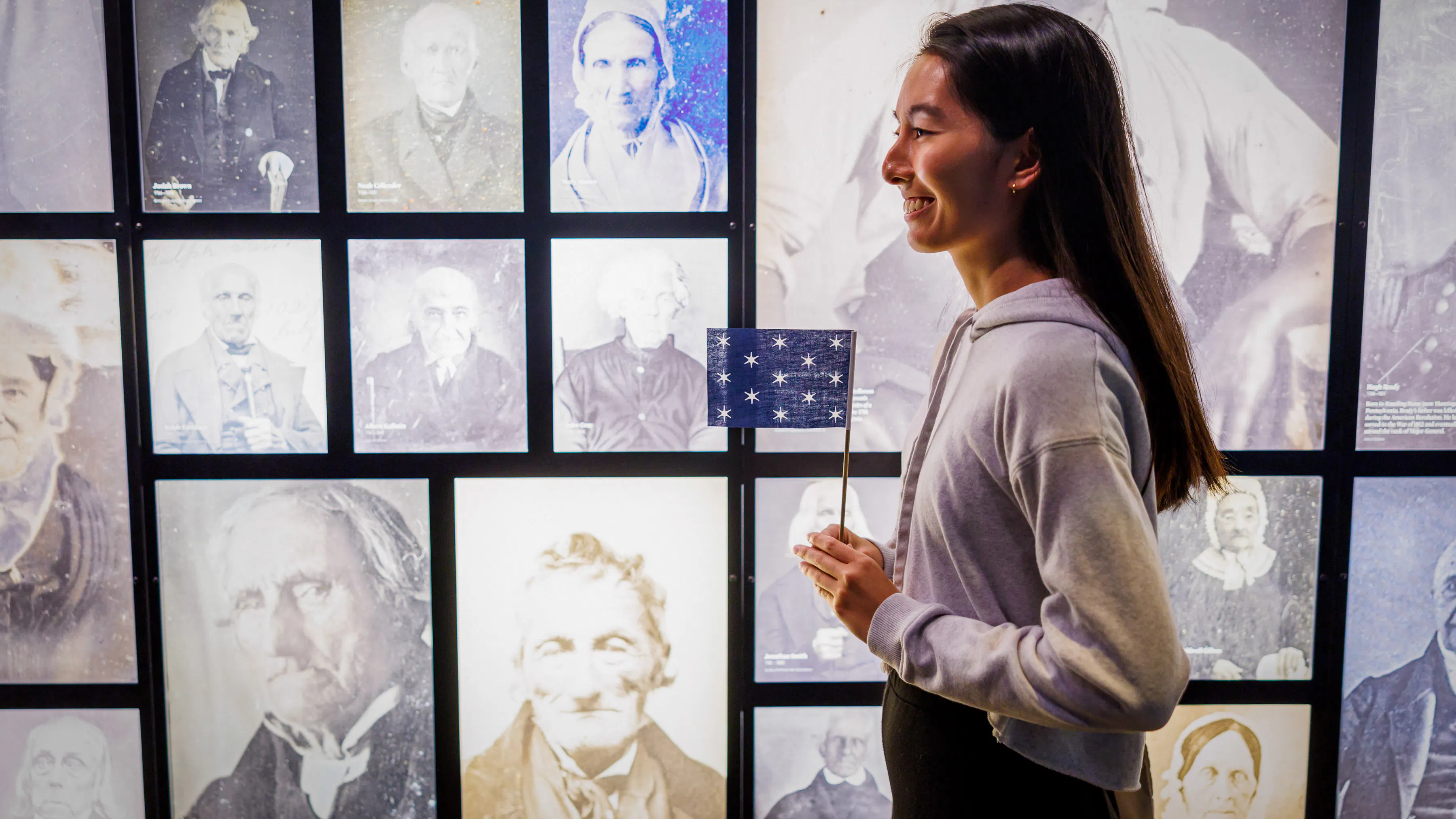 A visitor holds a miniature version of George Washington's standard flag in front of photographs of people from the Revolutionary generation who lived into the age of photography.