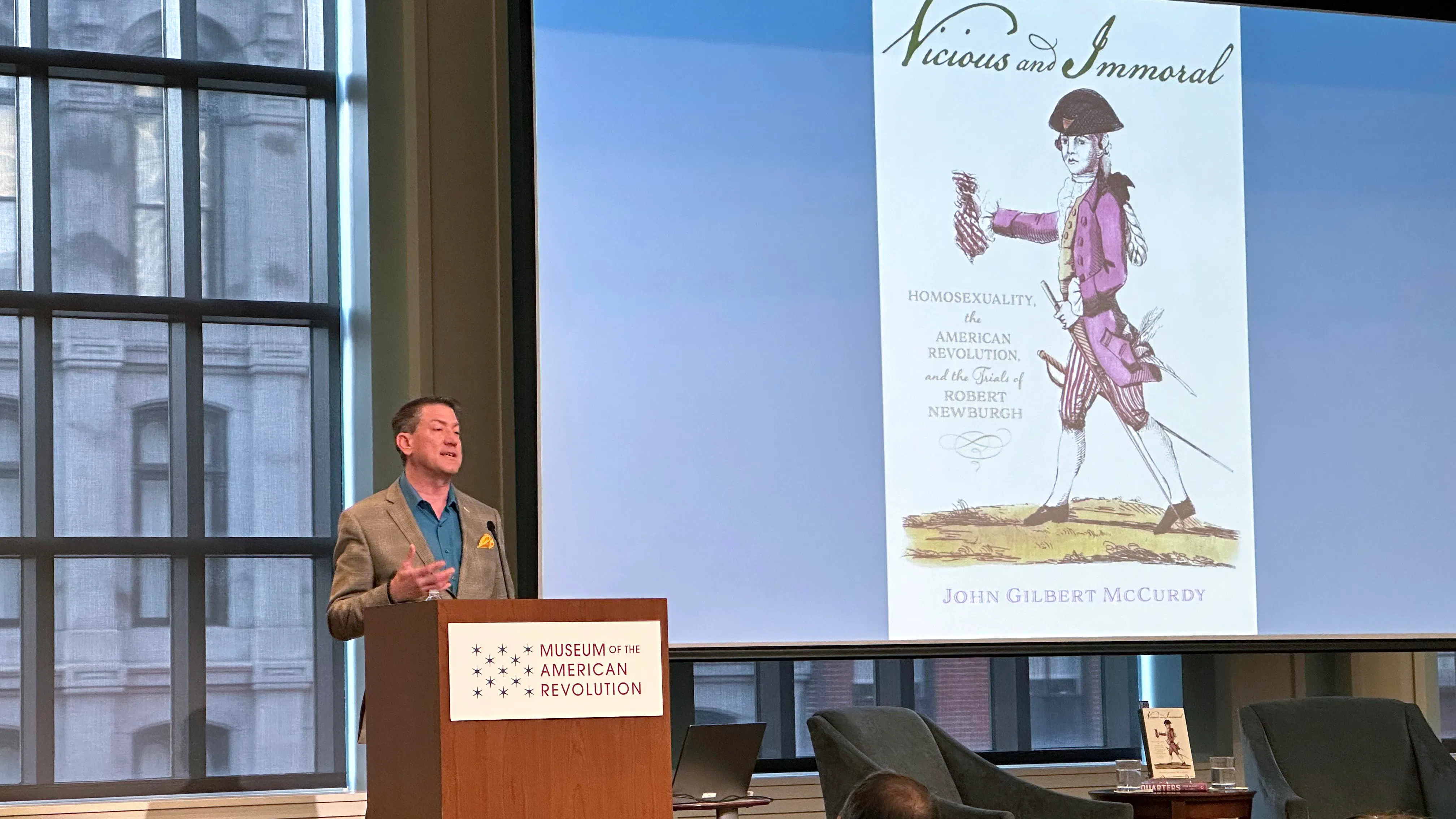 Dr. John Gilbert McCurdy stands at a podium with the Museum of the American Revolution logo and an image of his Vicious and Immoral book on the screen behind him.