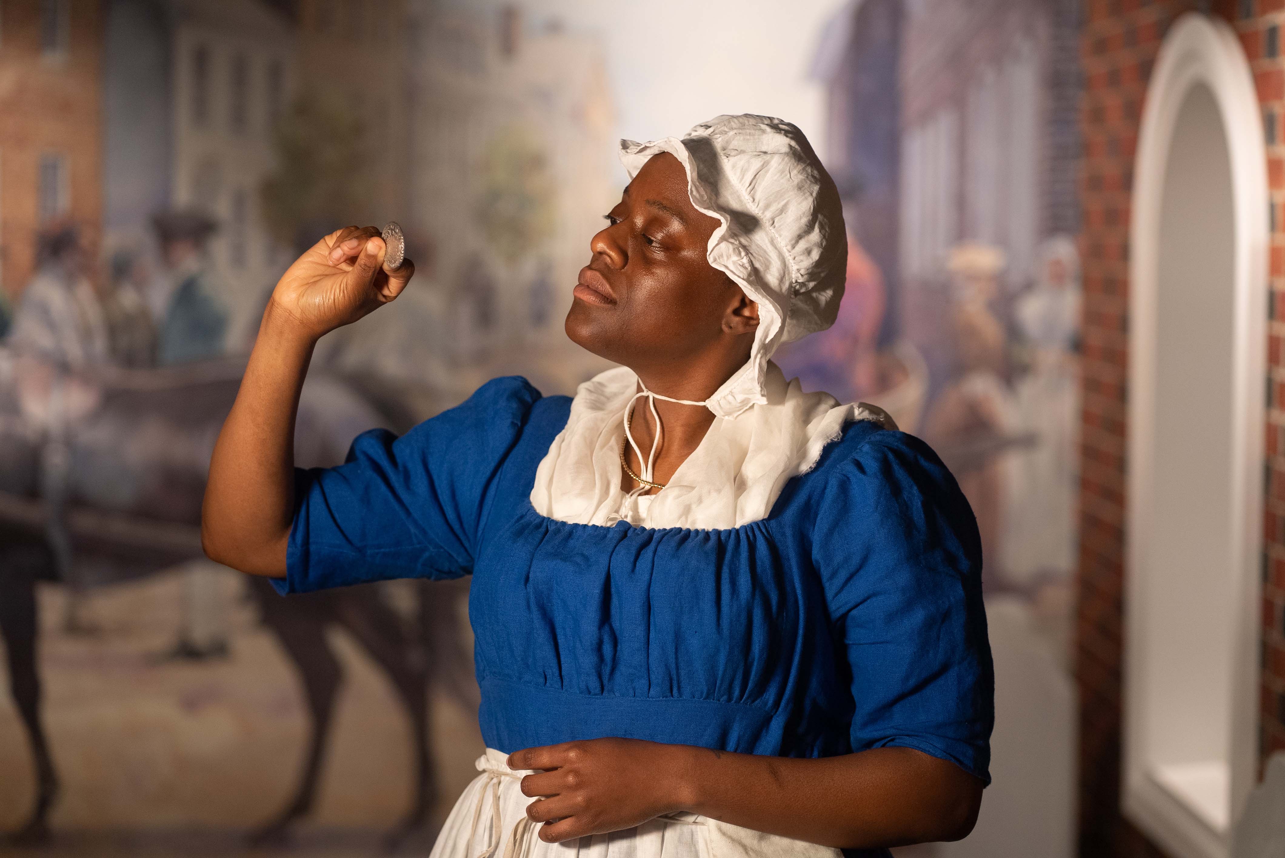 Katelyn Appiah-Kubi portrays Elizabeth Freeman wearing a blue dress with white apron and hat.