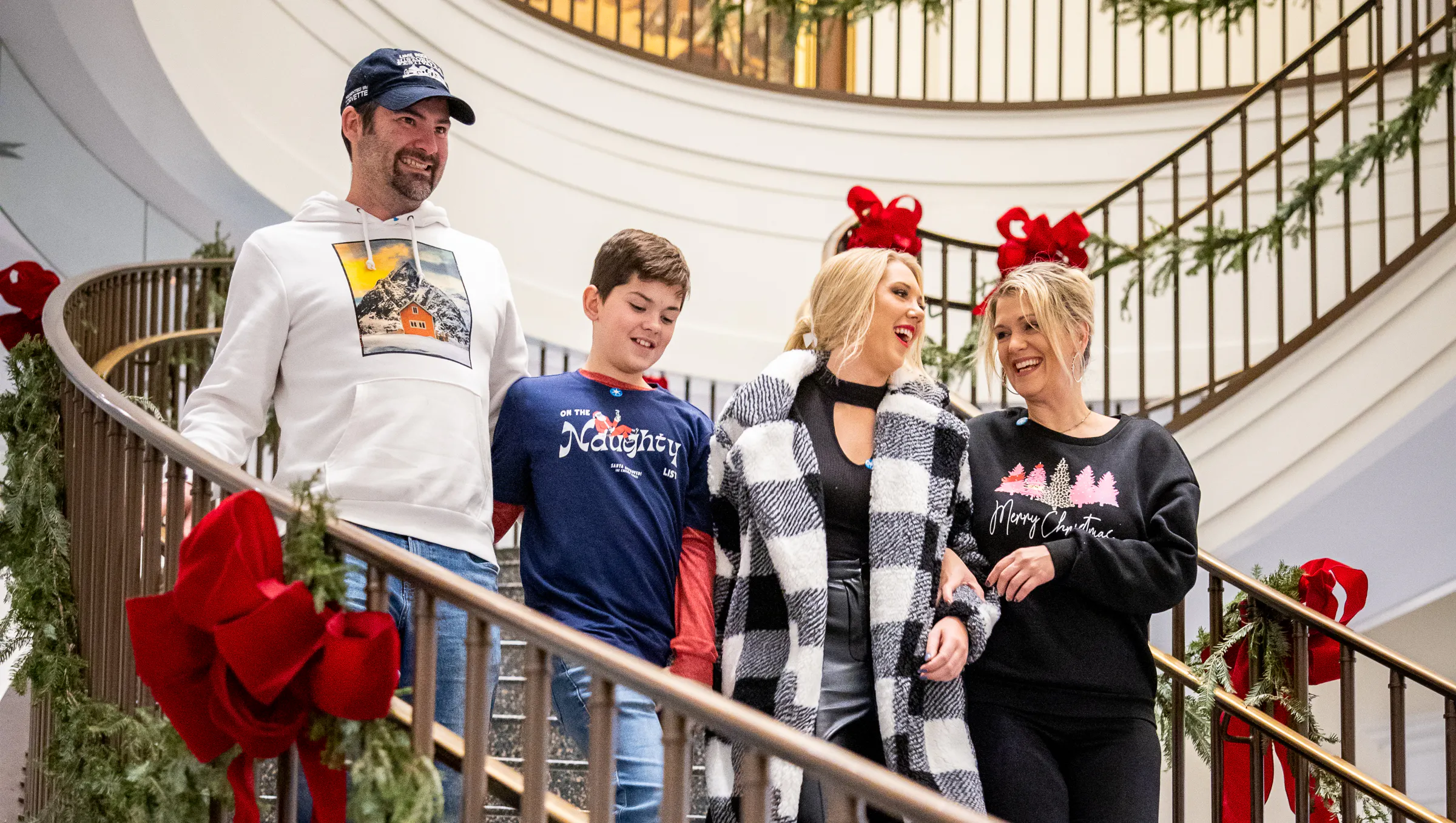 A family of four walks down the spiral staircase decorated with festive winter greenery and red bows.