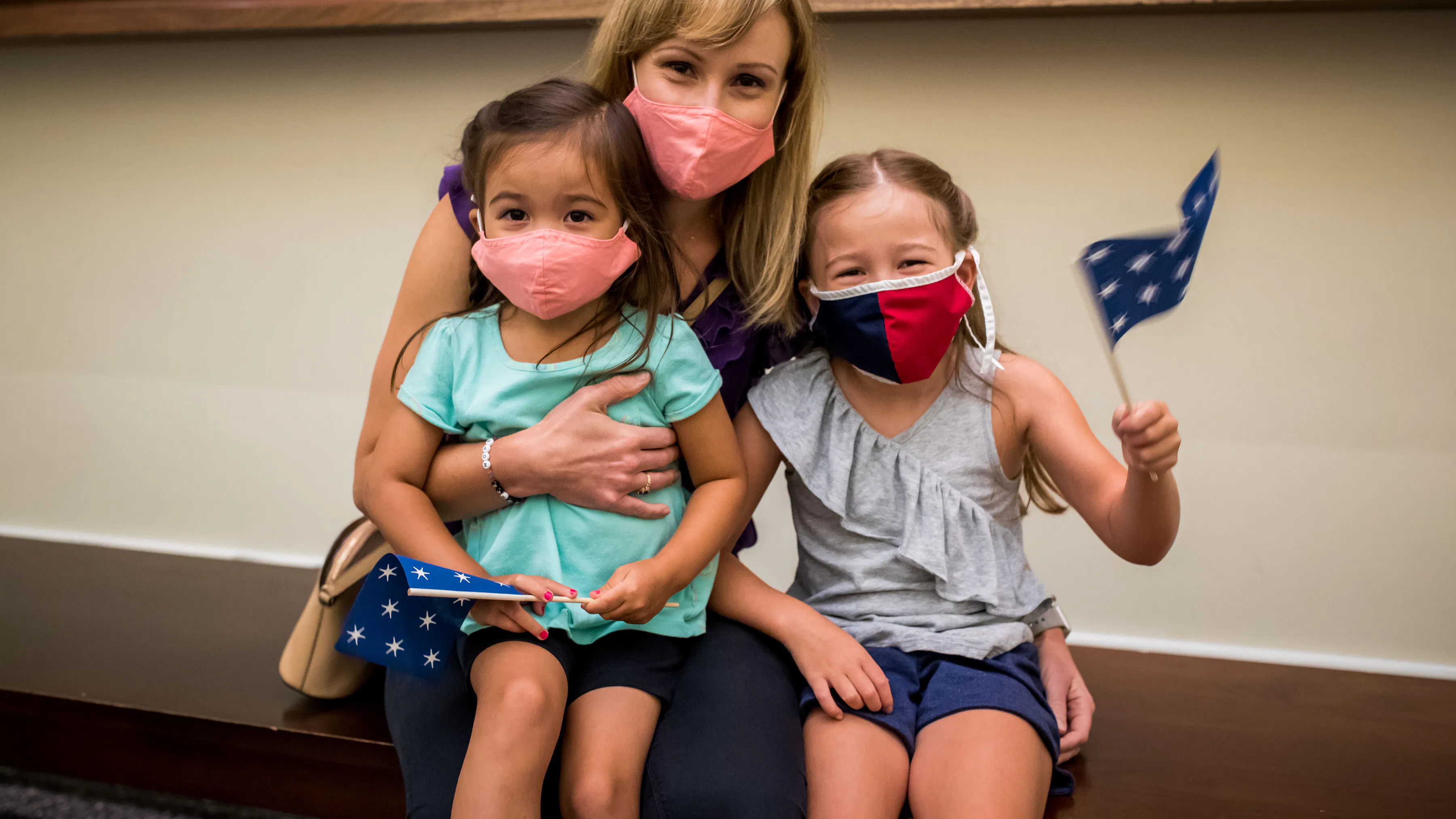 A mother and her two young daughters, all wearing masks, wave a 13-star flag while sitting on a bench in the Museum.