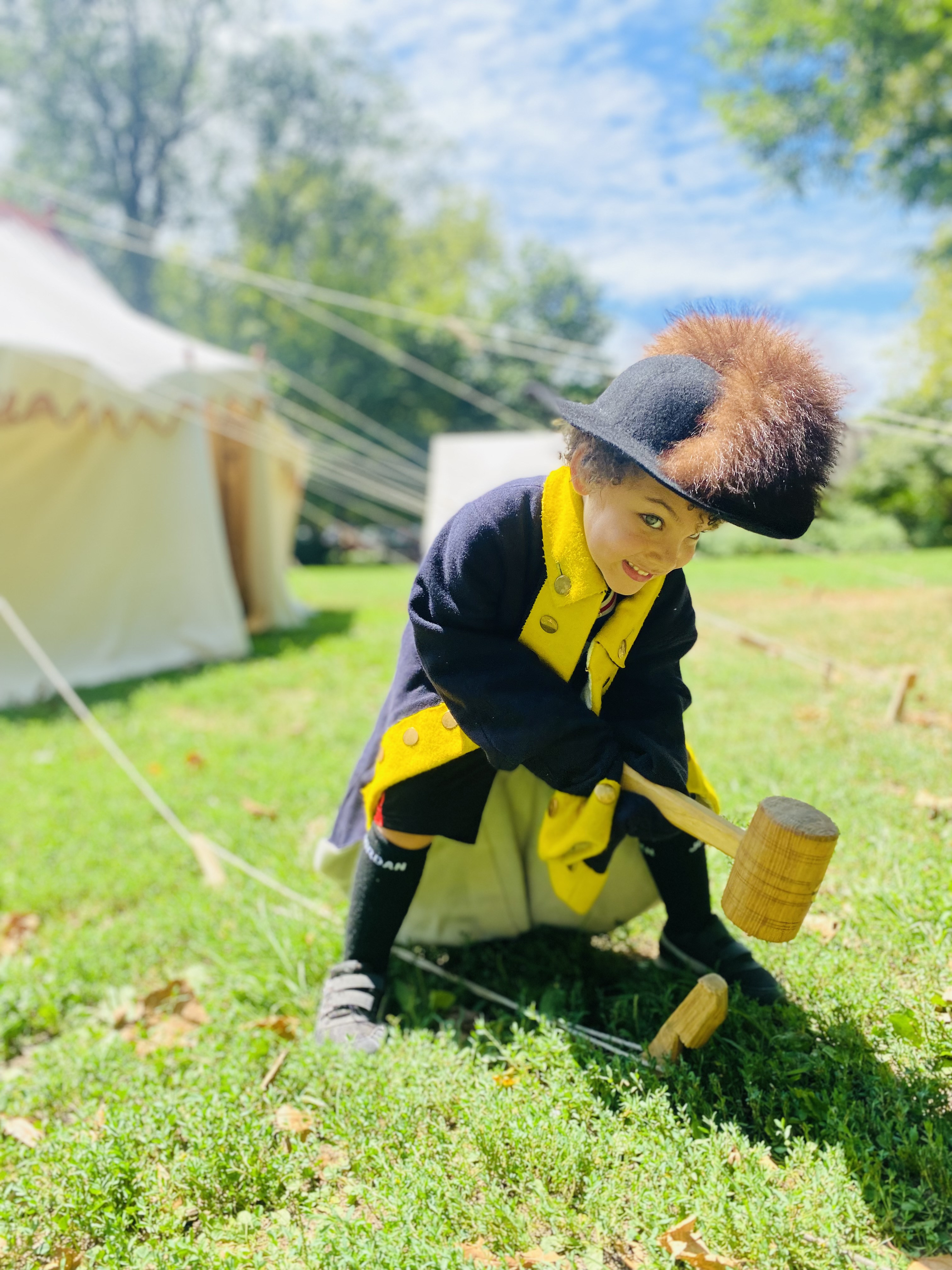 A child wearing replica clothing smiles while hammering a wooden stake for the Museum's First Oval office Project tent in Clark Park.
