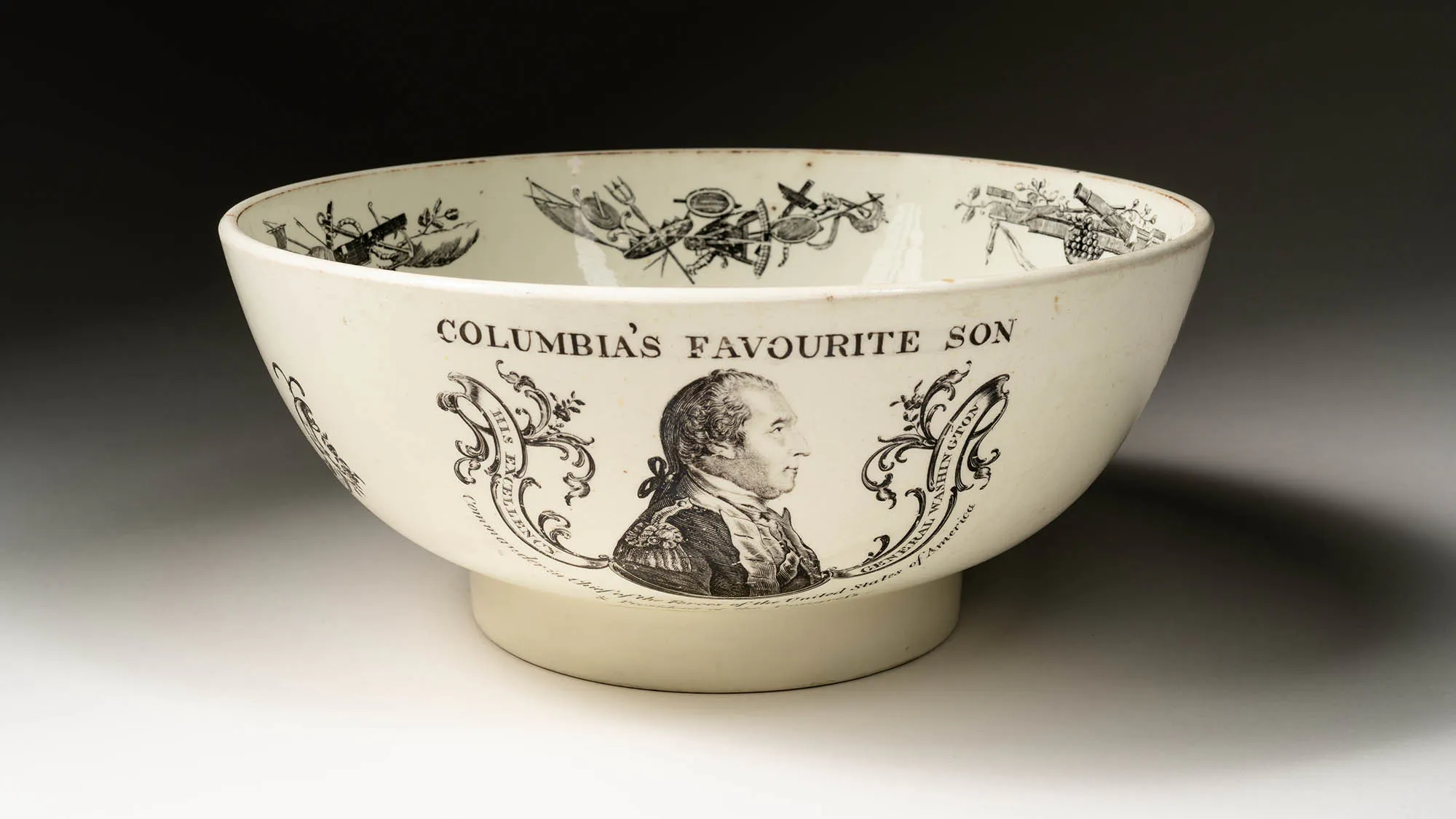 This image shows a Creamware Punchbowl. There is an engraving of a portrait of George Washington. He is seated facing the right. Above his image is text that reads “Columbia’s Favourite Son.” Inside the bowl are illustrations of cannons and swords.