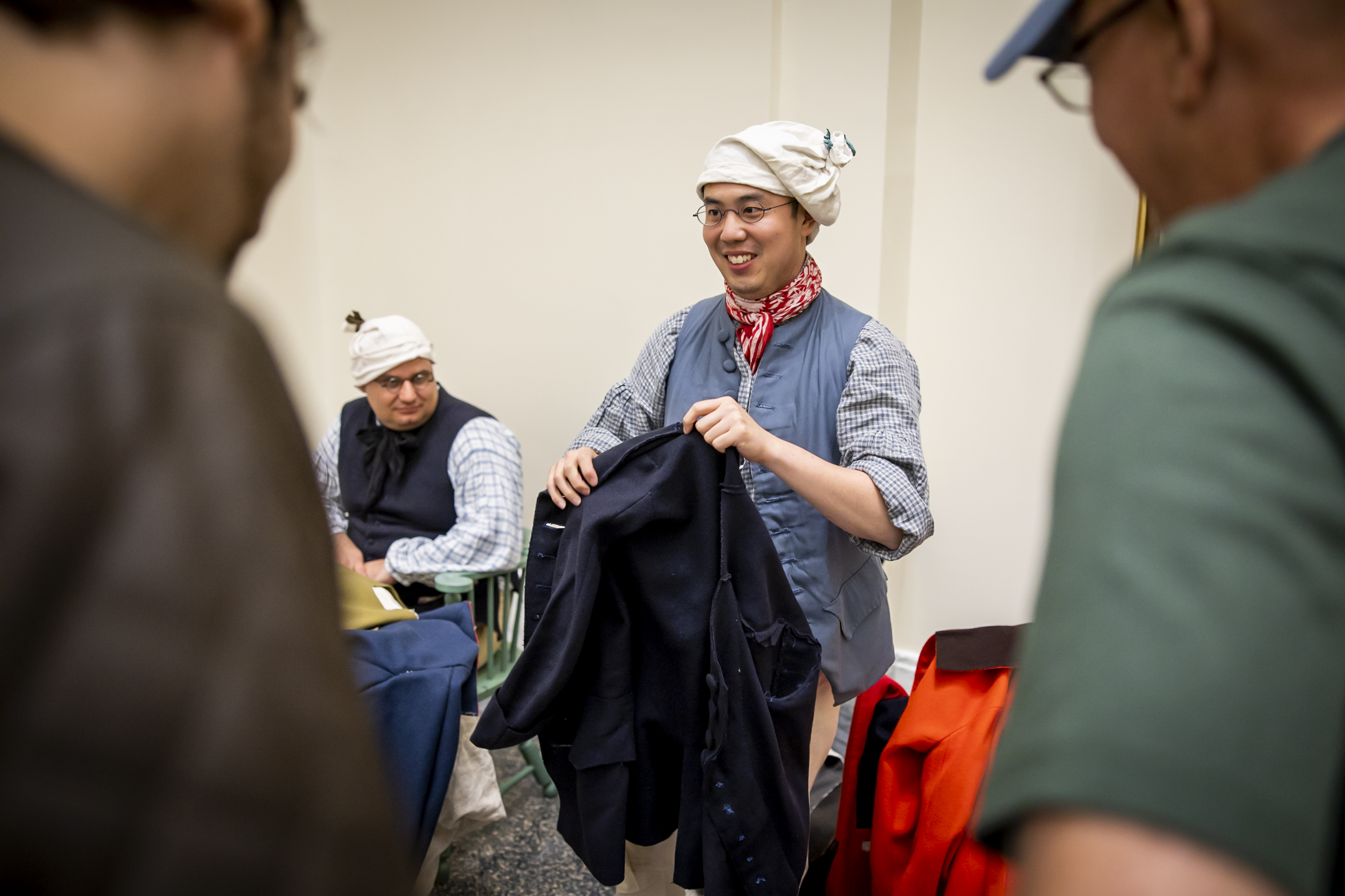 An Asian man dressed in 18th century clothing holds a piece of fabric. People stand in front of him, watching.