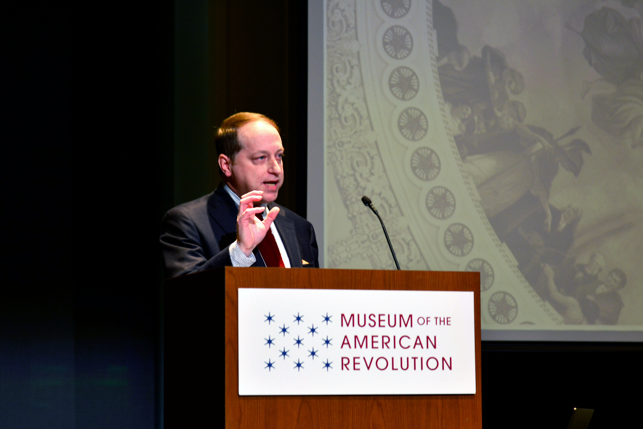 This image shows Michael Hattem standing at a wooden podium with the Museum's logo on the front while gesturing with his right hand