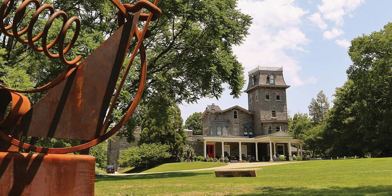 This image shows a sculpture in the foreground with a castle-looking building in the background that is the Woodmere Art Museum.