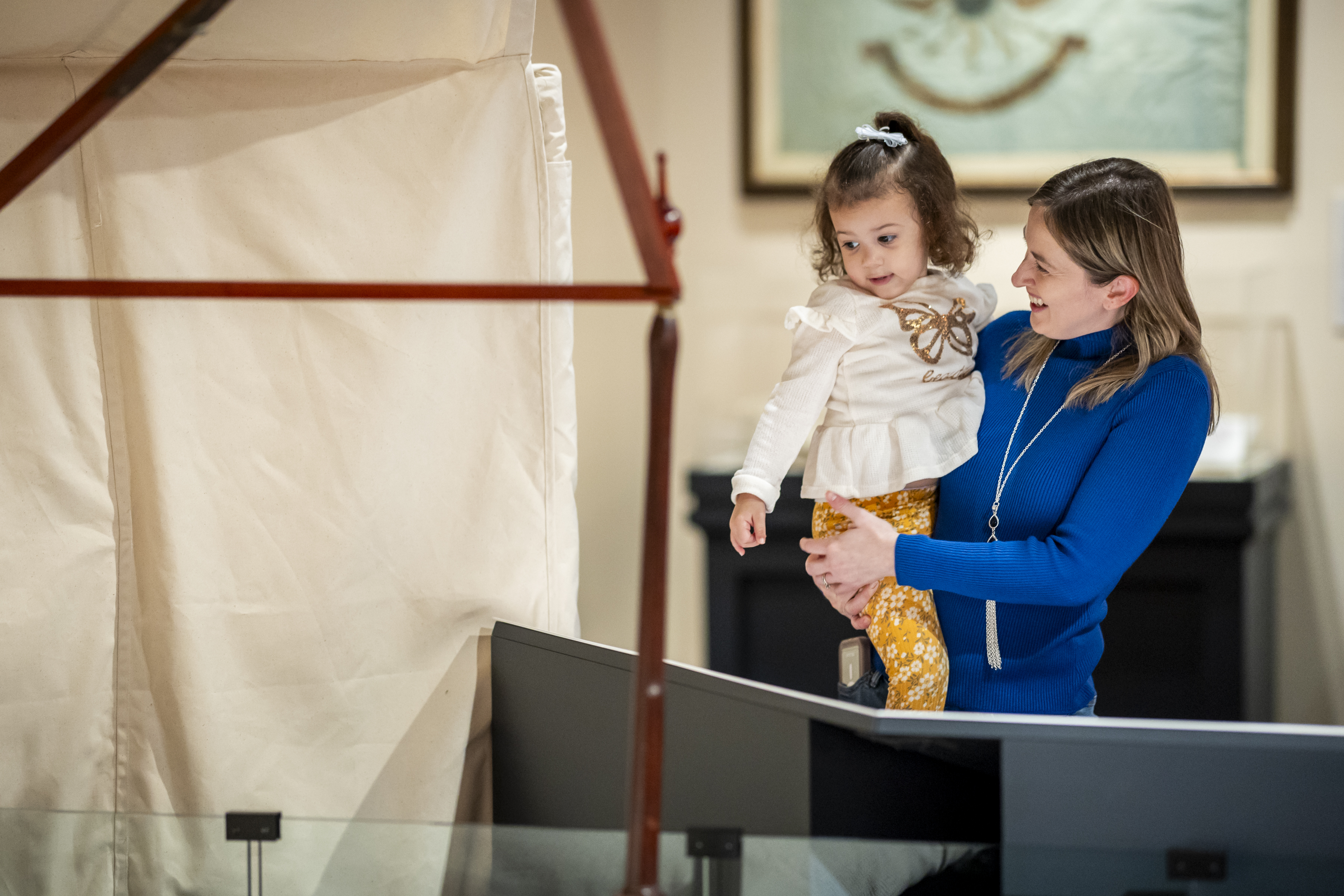 A mother holding her young daughter view Washington's camp bed on display in the Witness to Revolution exhibit.