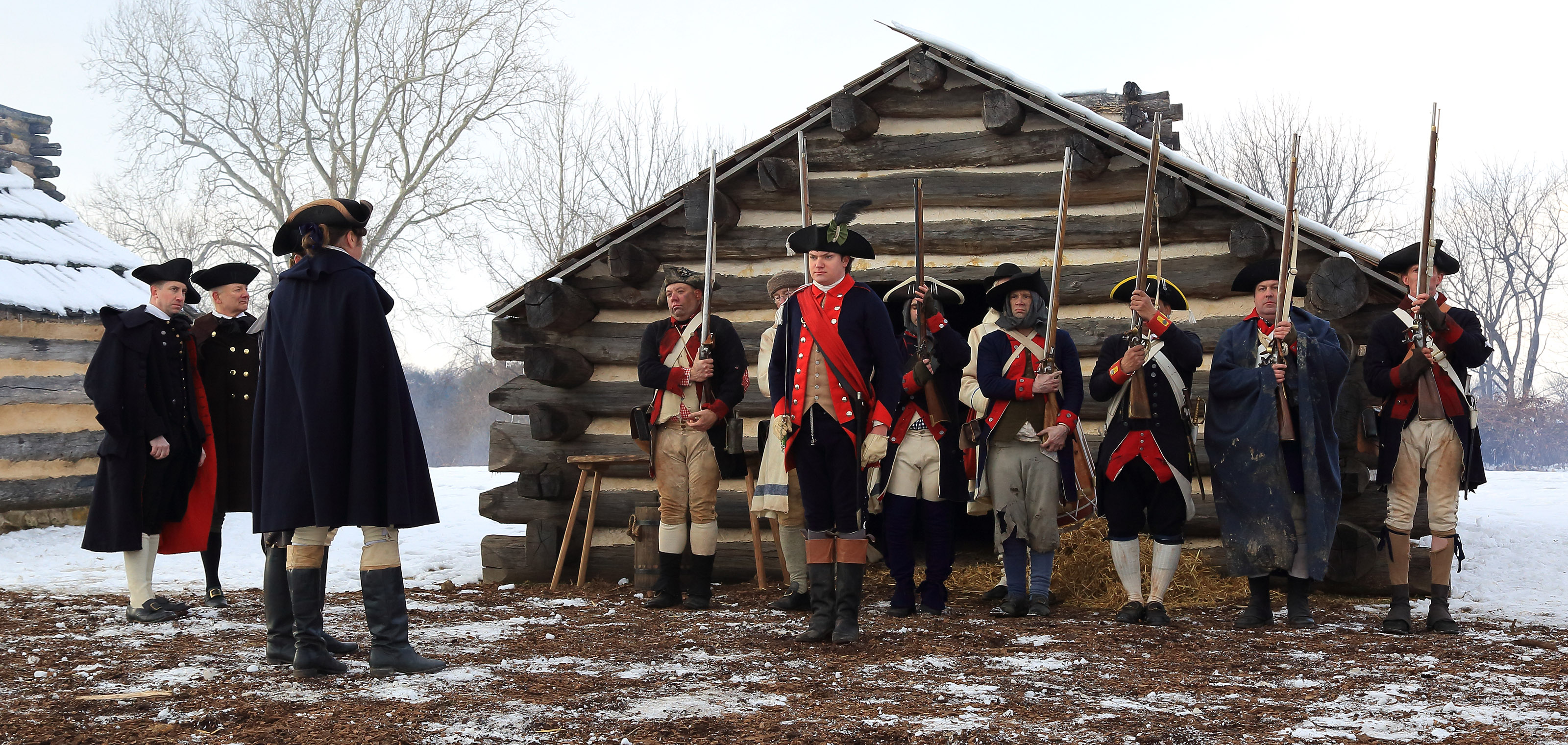 Costumed living historians dressed as the Continental Army stand in front of a log cabin at Valley Forge National Historical Park.