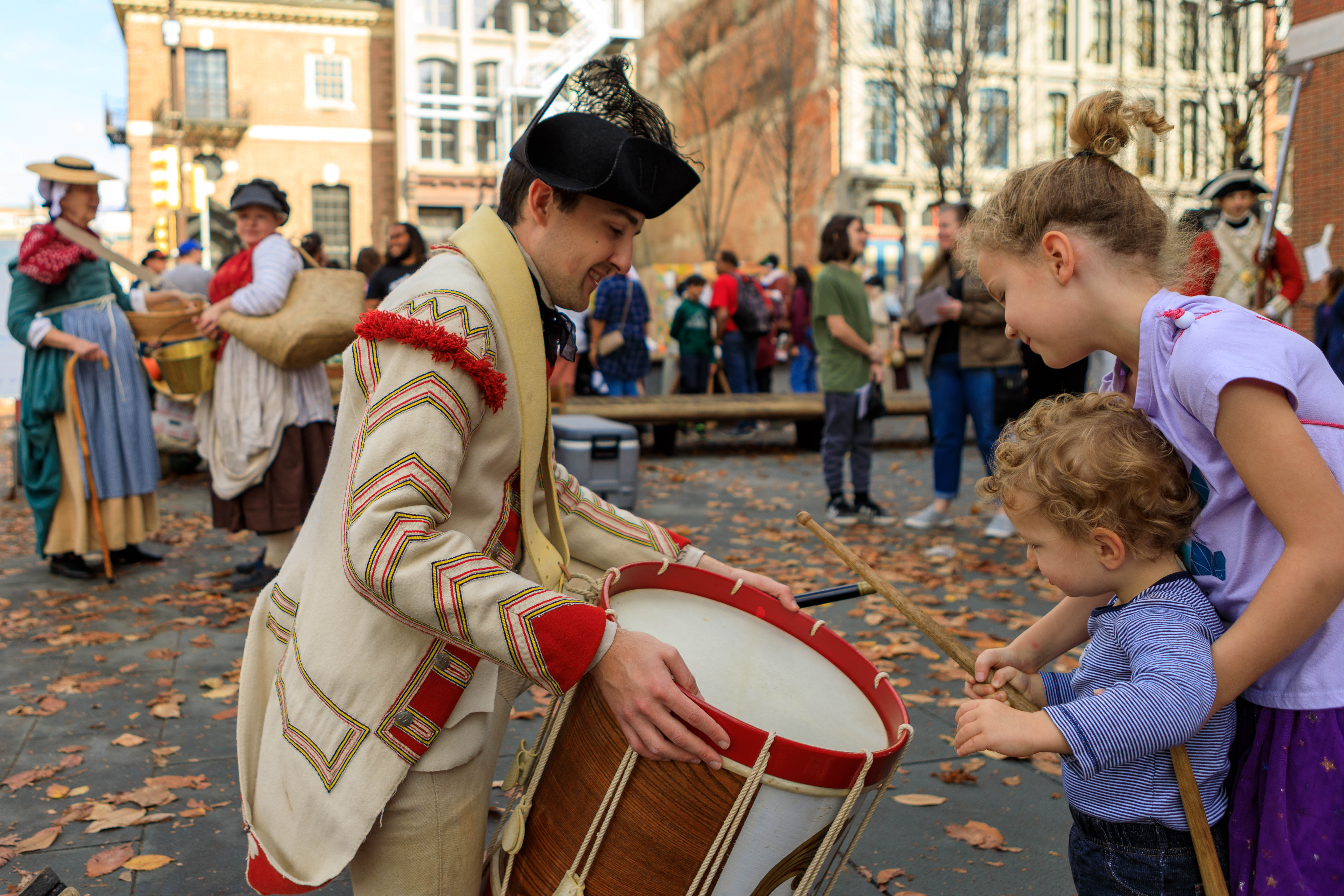 A costumed interpreter lets two young children play a drum on the Museum's plaza.