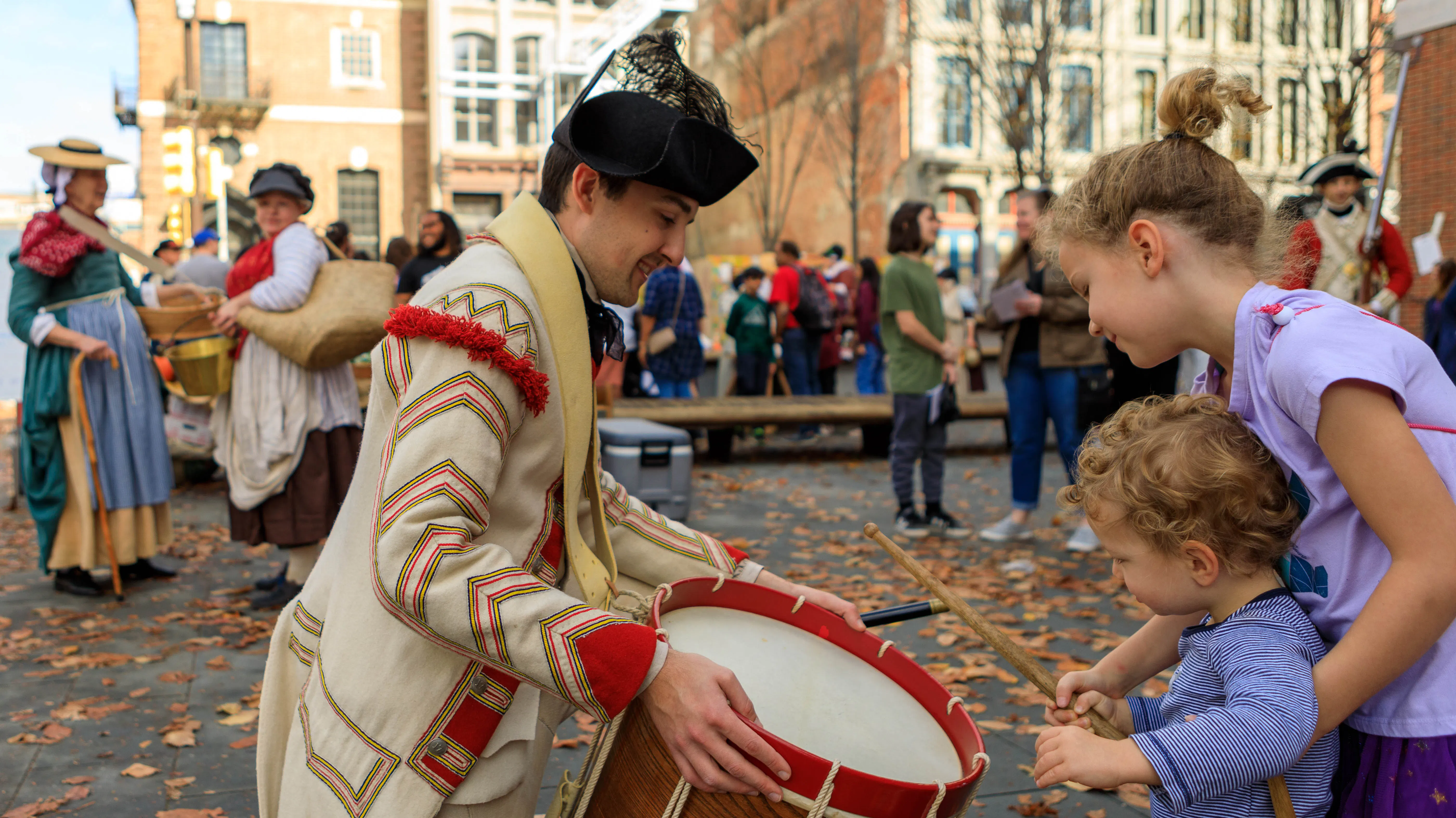A costumed interpreter lets two young children play a drum on the Museum's plaza.
