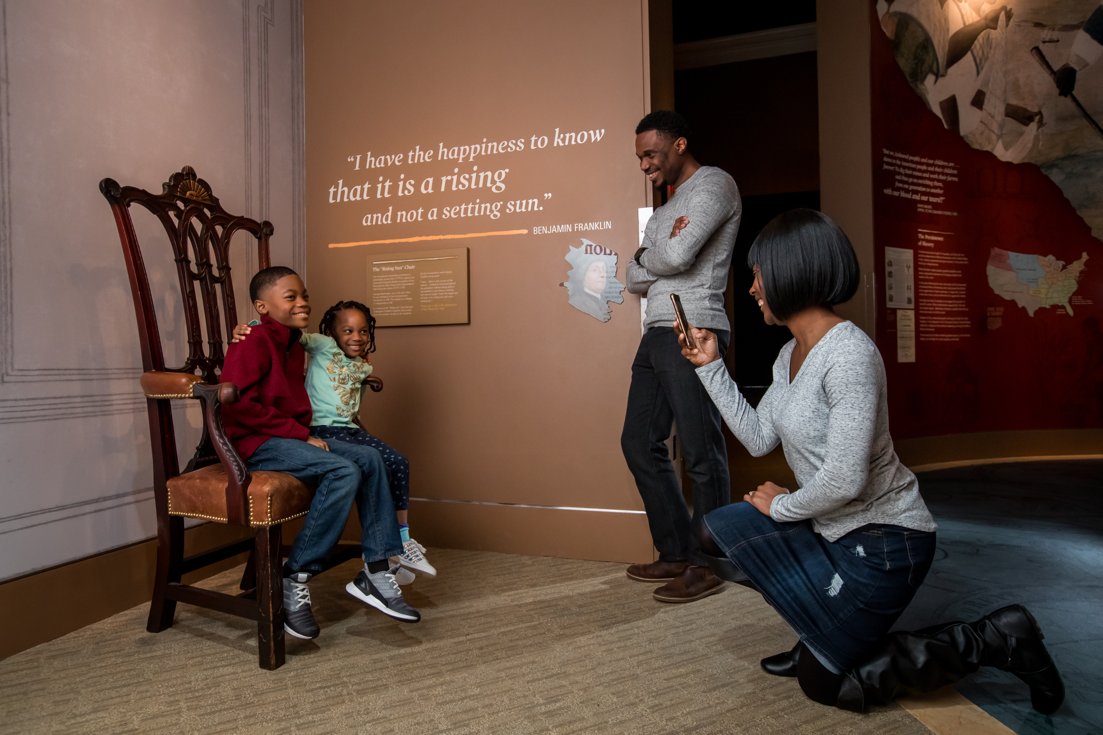 A mother takes a photo of her son and daughter sitting on a chair in the Writing the Constitution section inside the Museum galleries. She is bended on one knee as her husband stands next to her.
