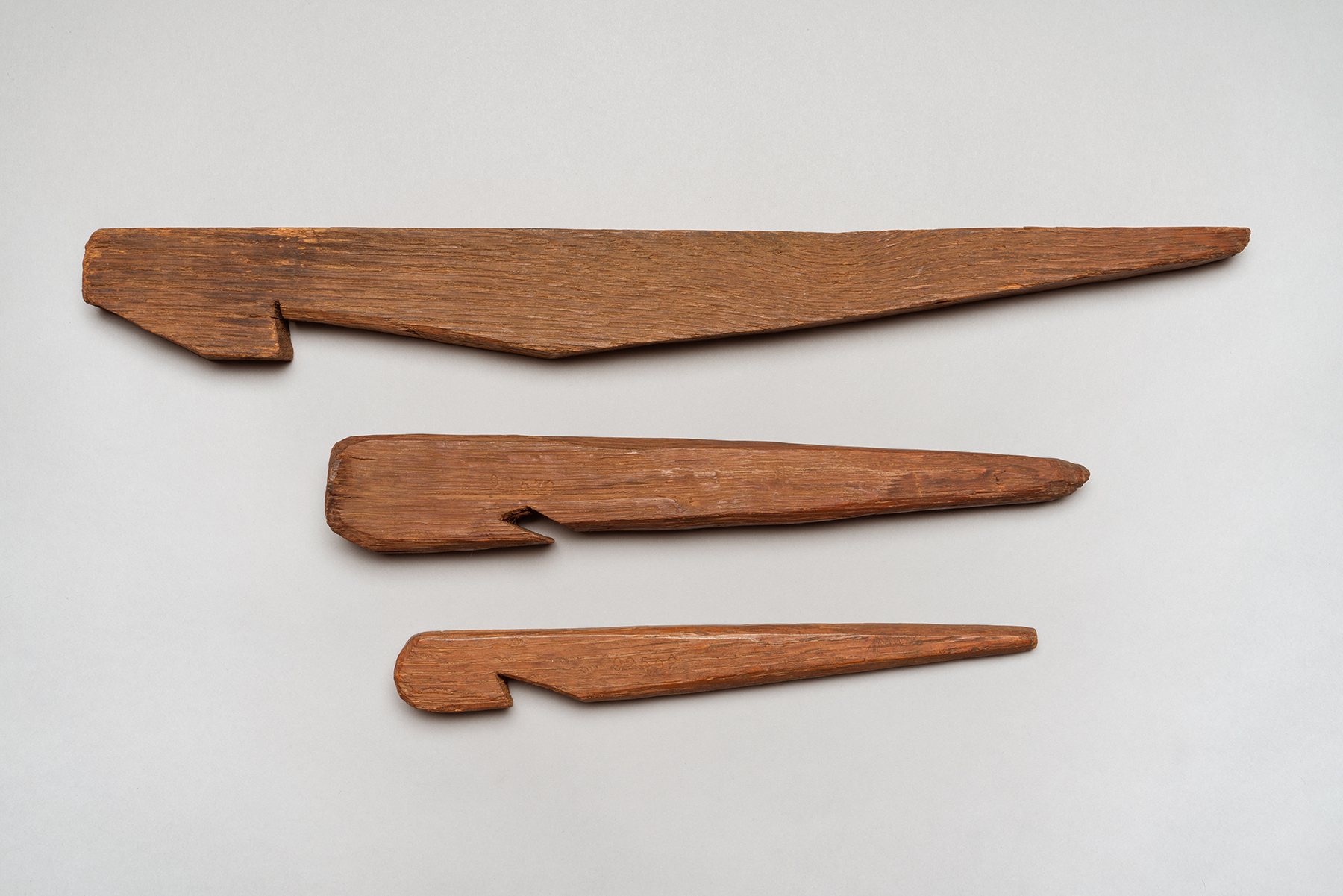 Three brown wooden tent stakes with one sharp end to dig into the ground and one notched end to hold rope.