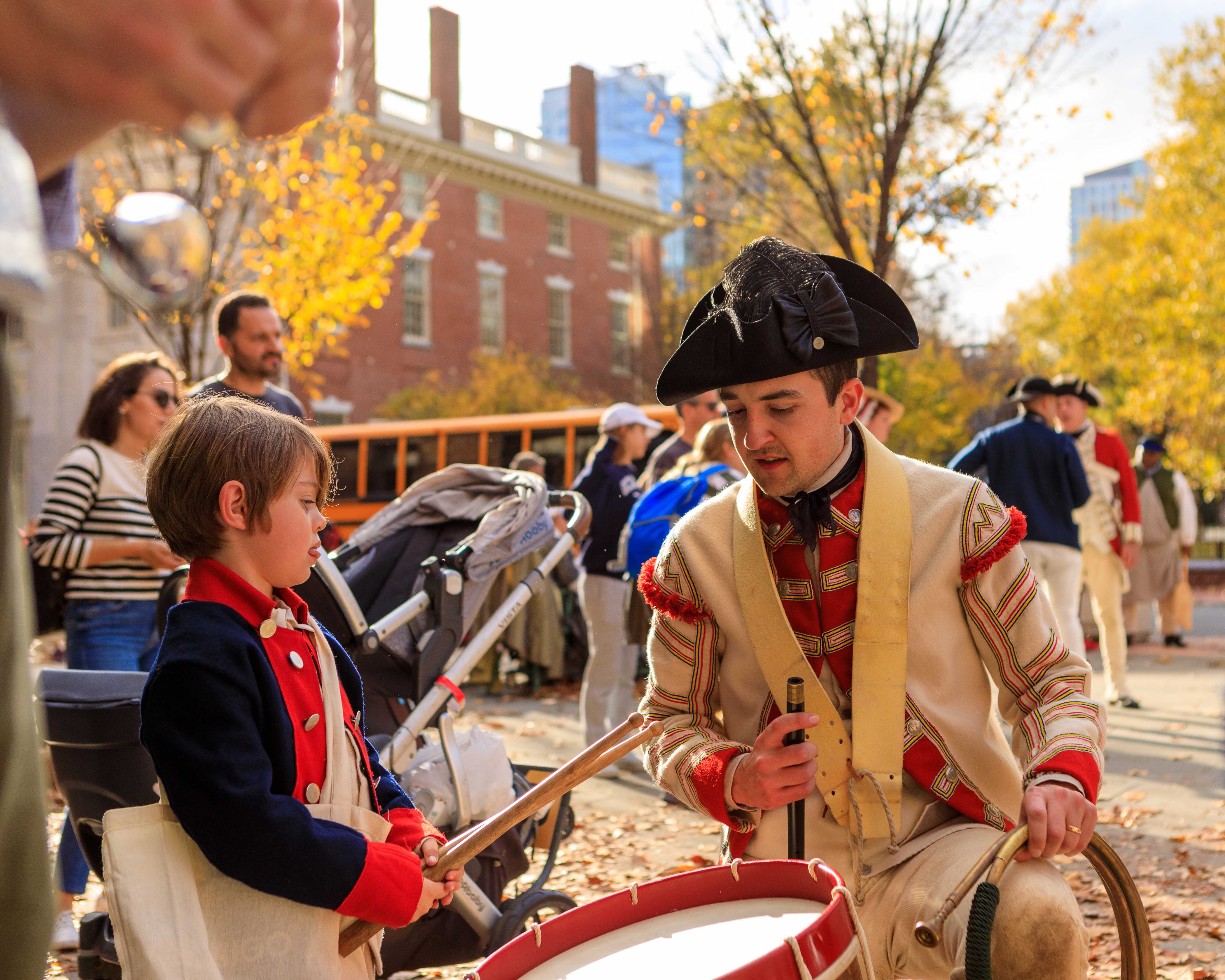 A British costumed living history interpreter shows a young boy how to drum on the Museum's plaza during Occupied Philadelphia.